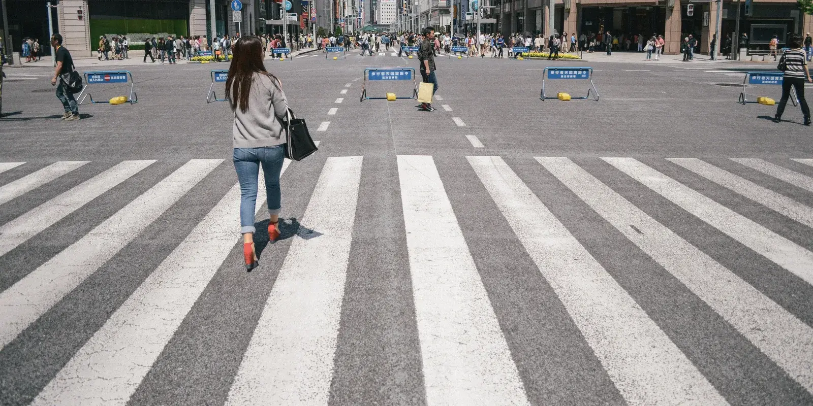A wide pedestrian crossing at the Ginza 4-chome intersection in Tokyo, with a person walking away across the stripes and others moving at a distance under bright daylight