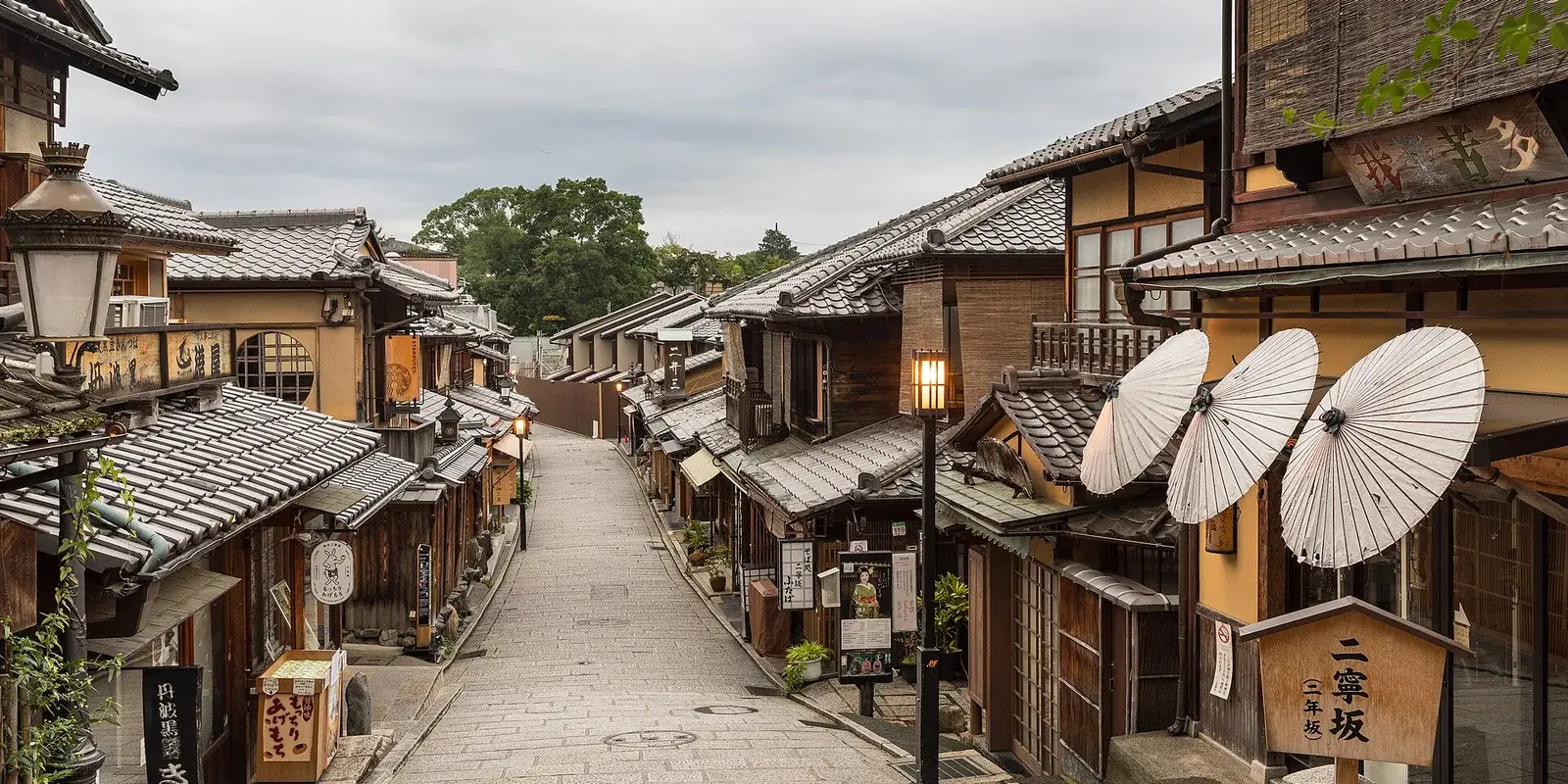 A quiet early-morning view down Ninenzaka, a sloping cobbled lane of traditional wooden shops with paper umbrellas in Higashiyama, Kyoto, with no people in sight