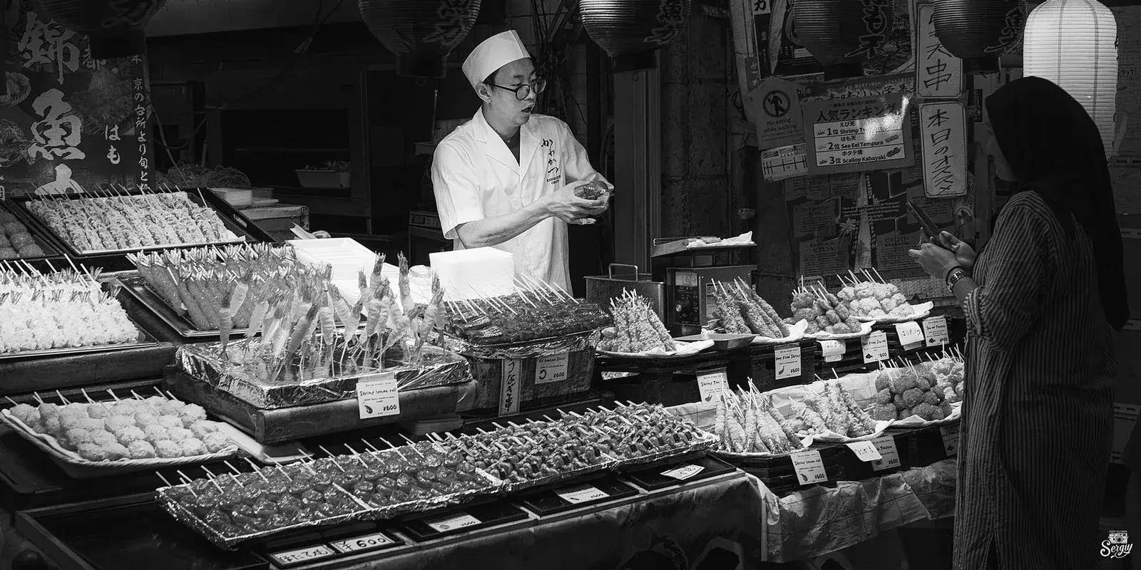 A merchant at Kyoto's Nishiki Market offers a skewered snack to a customer across his food stall lined with lanterns
