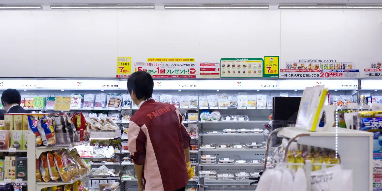 Interior of a Japanese 7-Eleven convenience store in Kyoto, with chilled shelves of bento and sandwiches and a staff member seen from behind