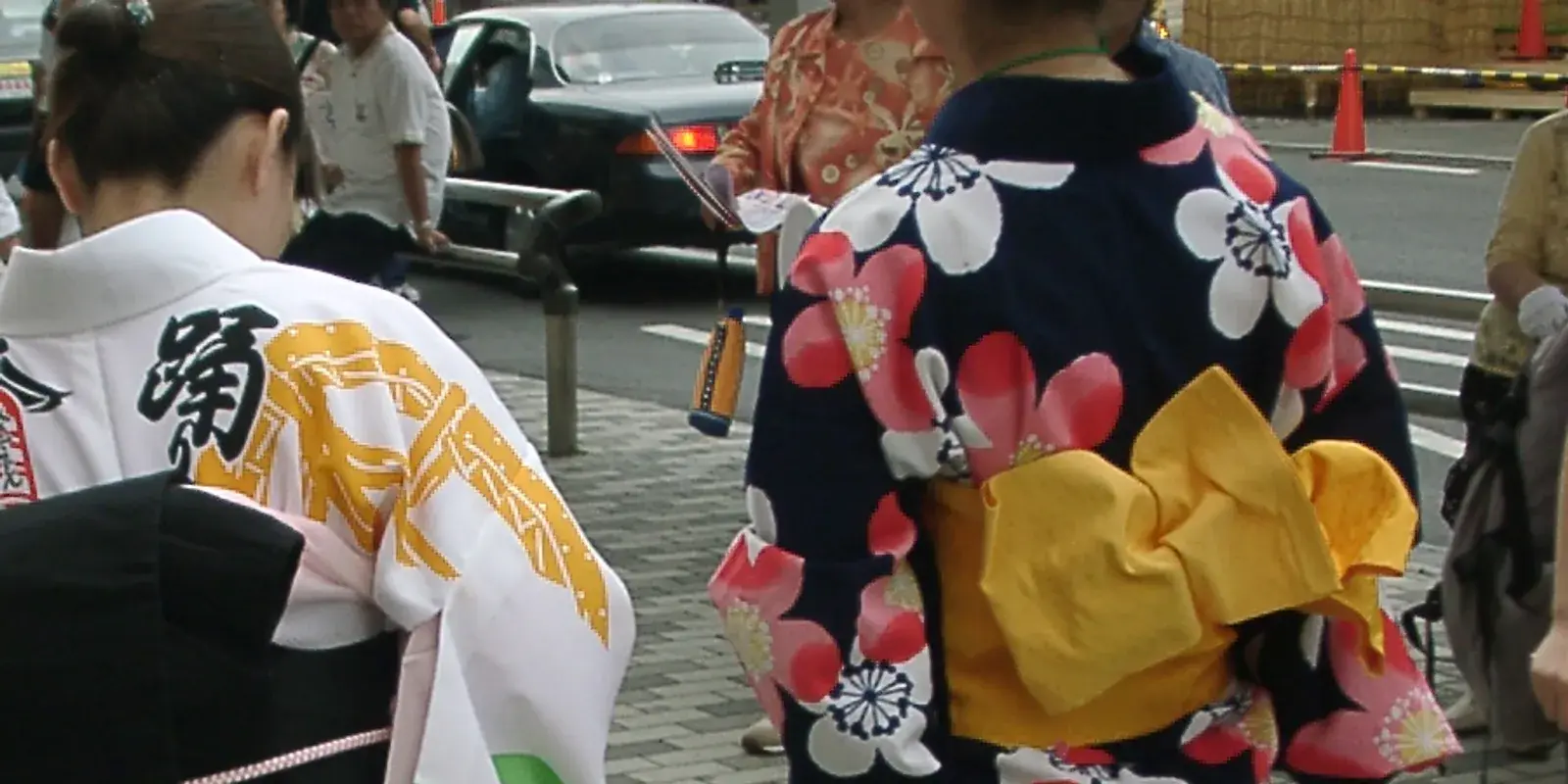 Two people seen from behind on a sunny festival street in Japan, one wearing a navy yukata patterned with pink and white blossoms tied with a bright yellow obi bow, the other in a white and gold happi coat, with no faces visible