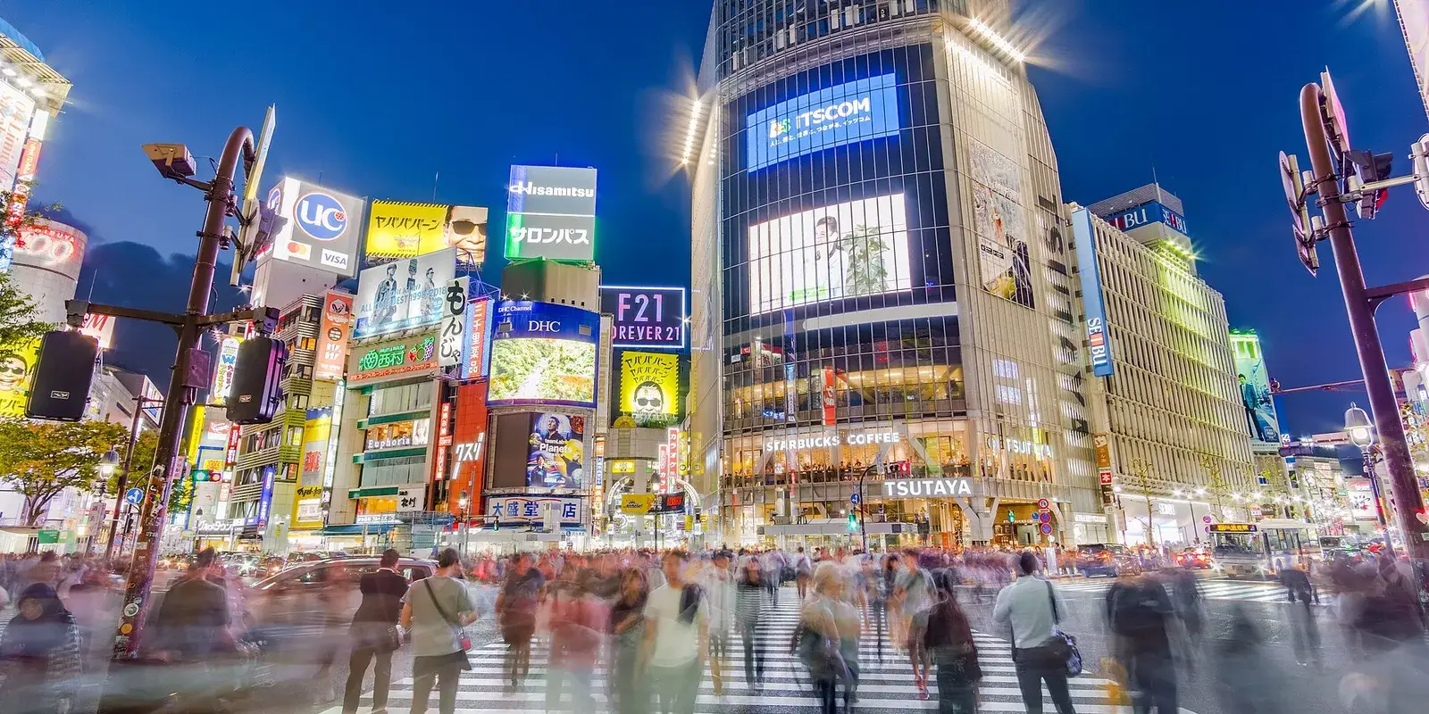 Crowds cross the Shibuya Scramble Crossing in Tokyo at dusk, the pedestrians blurred in motion beneath glowing neon billboards