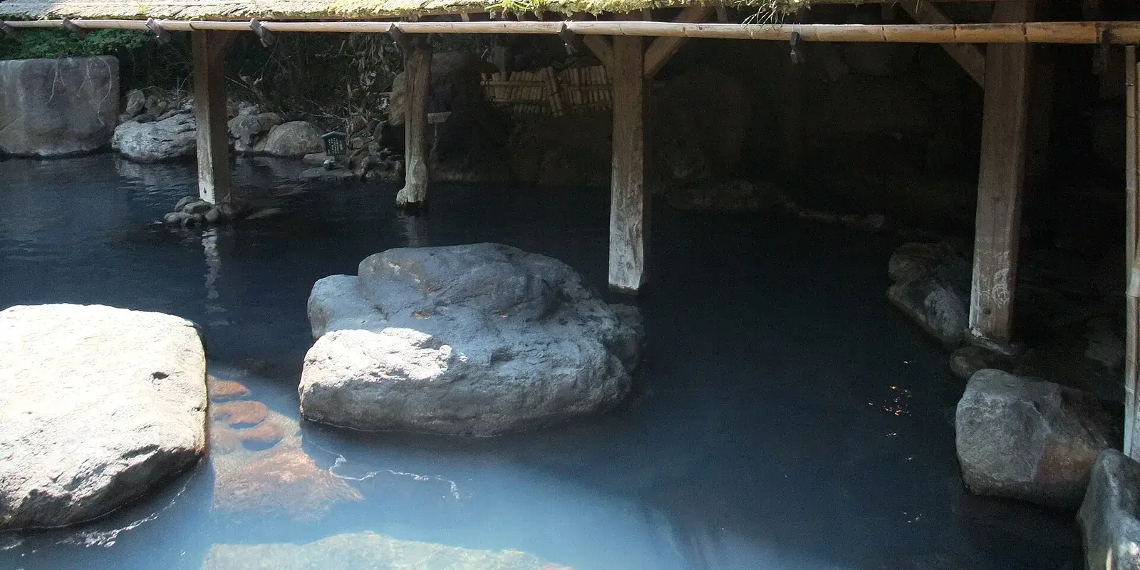 Empty outdoor onsen (rotenburo) at Kurokawa Onsen, Kyushu, with rocks in steaming milky water beneath a thatched-roof shelter