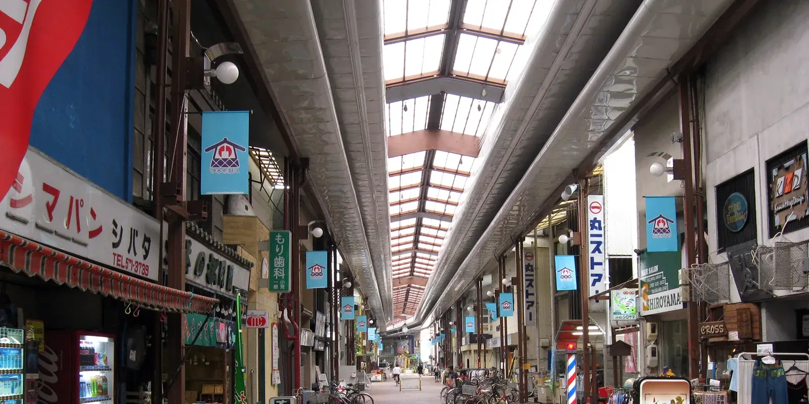 A quiet covered shopping arcade (shotengai) in Nagoya, Japan, with everyday local shops lining both sides, parked bicycles, and a skylit roof leading the eye down the empty street