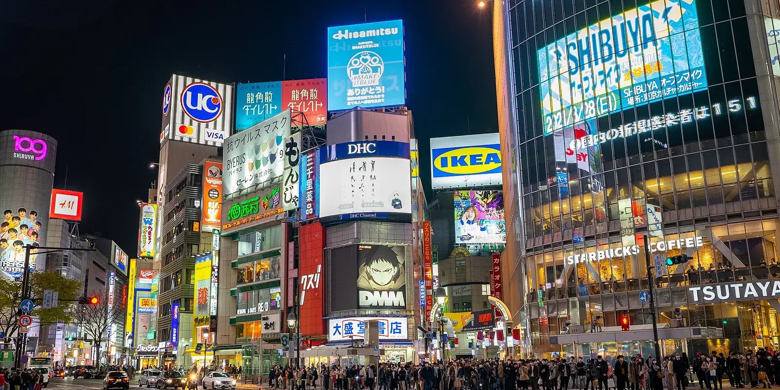 The Shibuya scramble crossing in Tokyo at night, lit by neon billboards, with a distant crowd of people streaming across the intersection