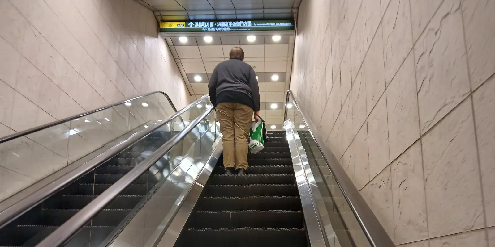 A commuter stands still while riding up an escalator at a Tokyo subway station, seen from behind
