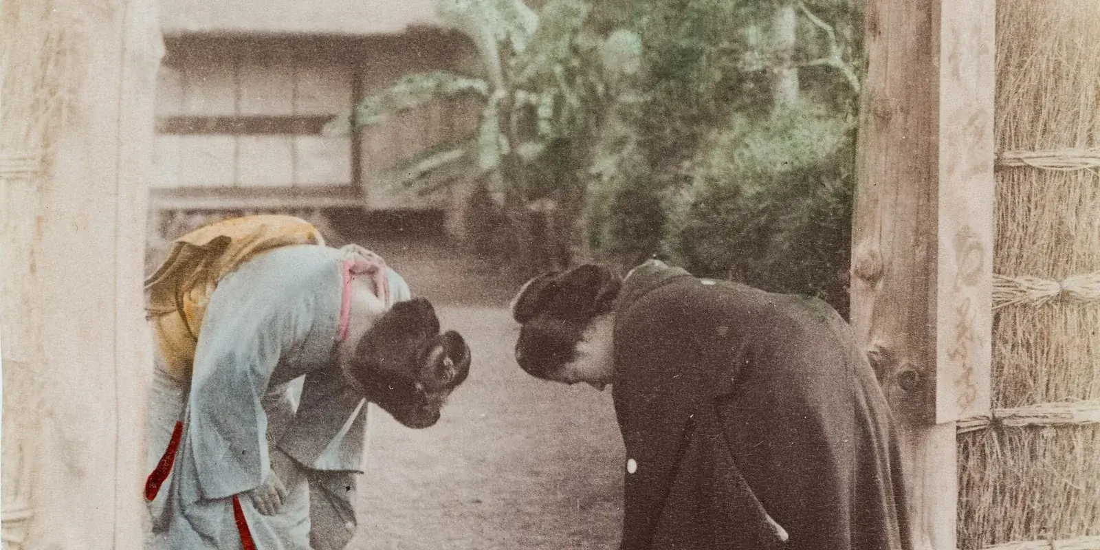 Two women in kimono bowing to each other (ojigi) under a thatched gate in a Japanese garden, hand-colored photograph circa 1880