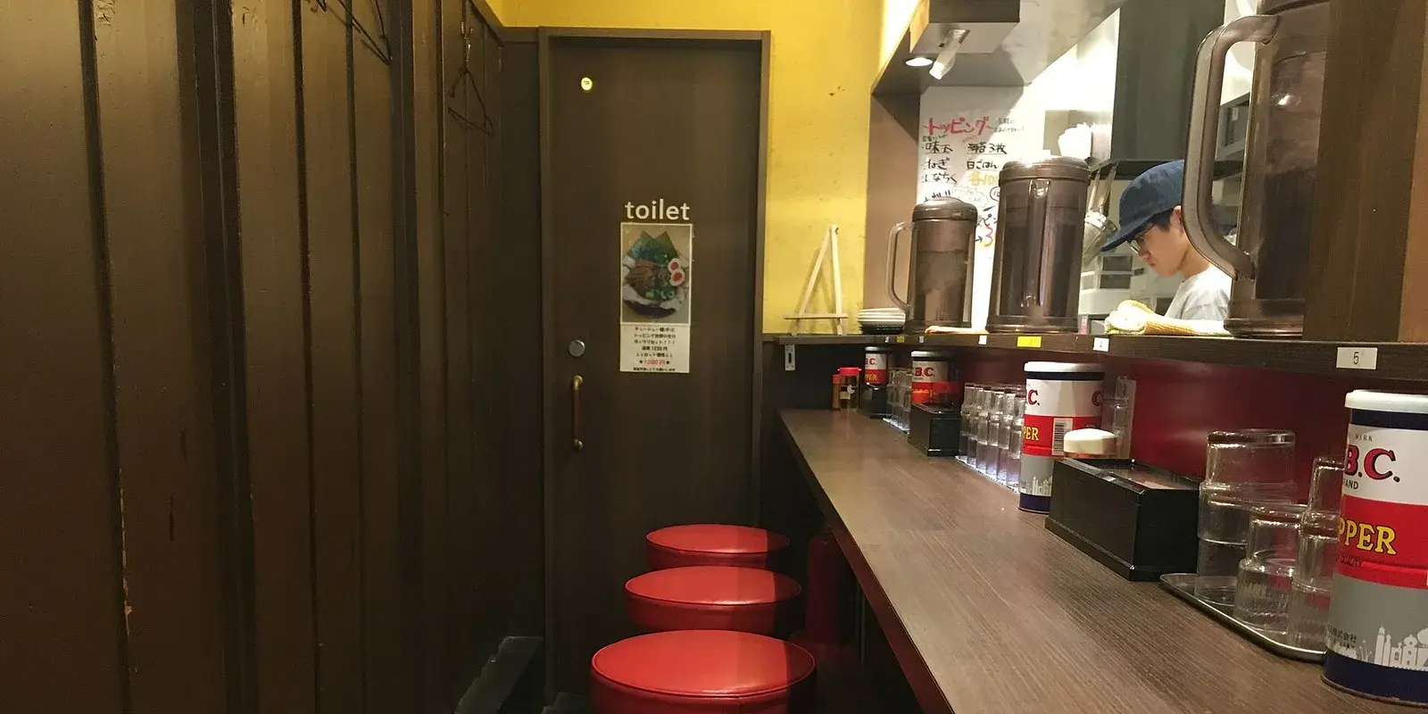 Row of single red counter stools along a wooden ramen-shop counter, the kind of seat built for one person