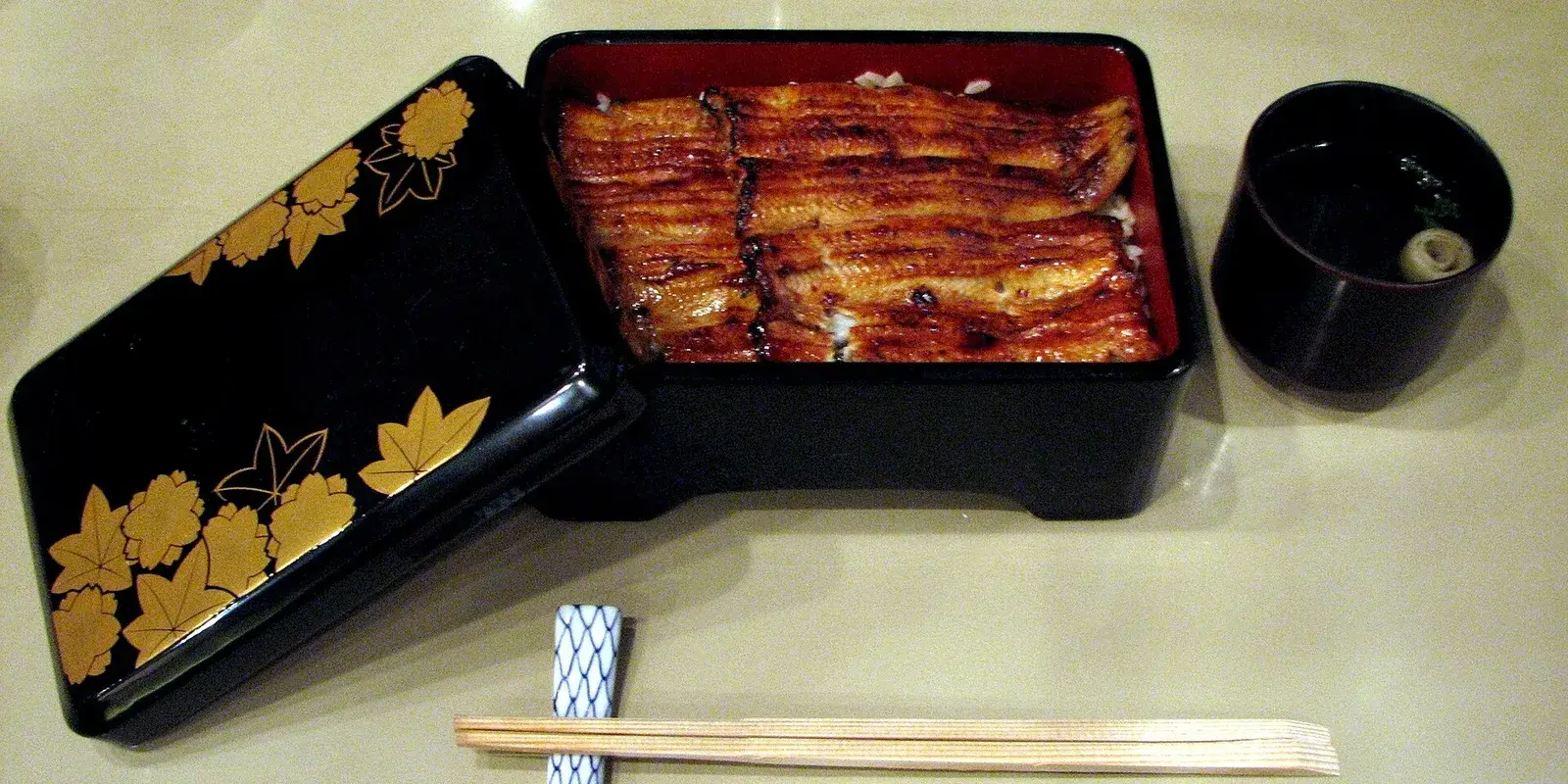 Open lacquer eel box (unaju) with glossy grilled eel over rice, a gold maple-leaf lid, chopsticks, and a soup cup