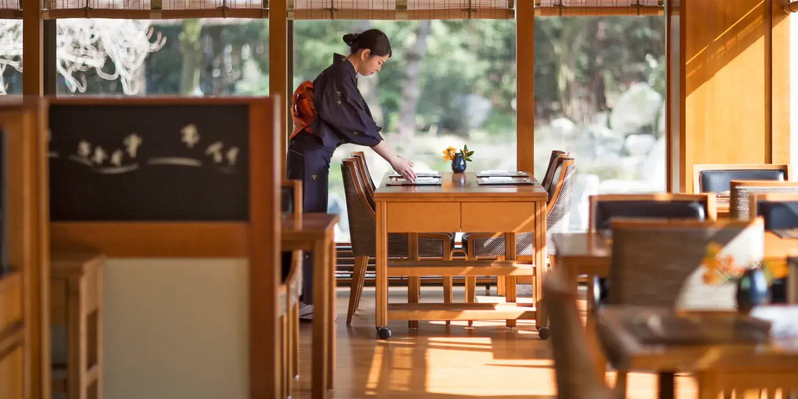 A waitress in a kimono carefully sets the table in a quiet Japanese restaurant overlooking a garden