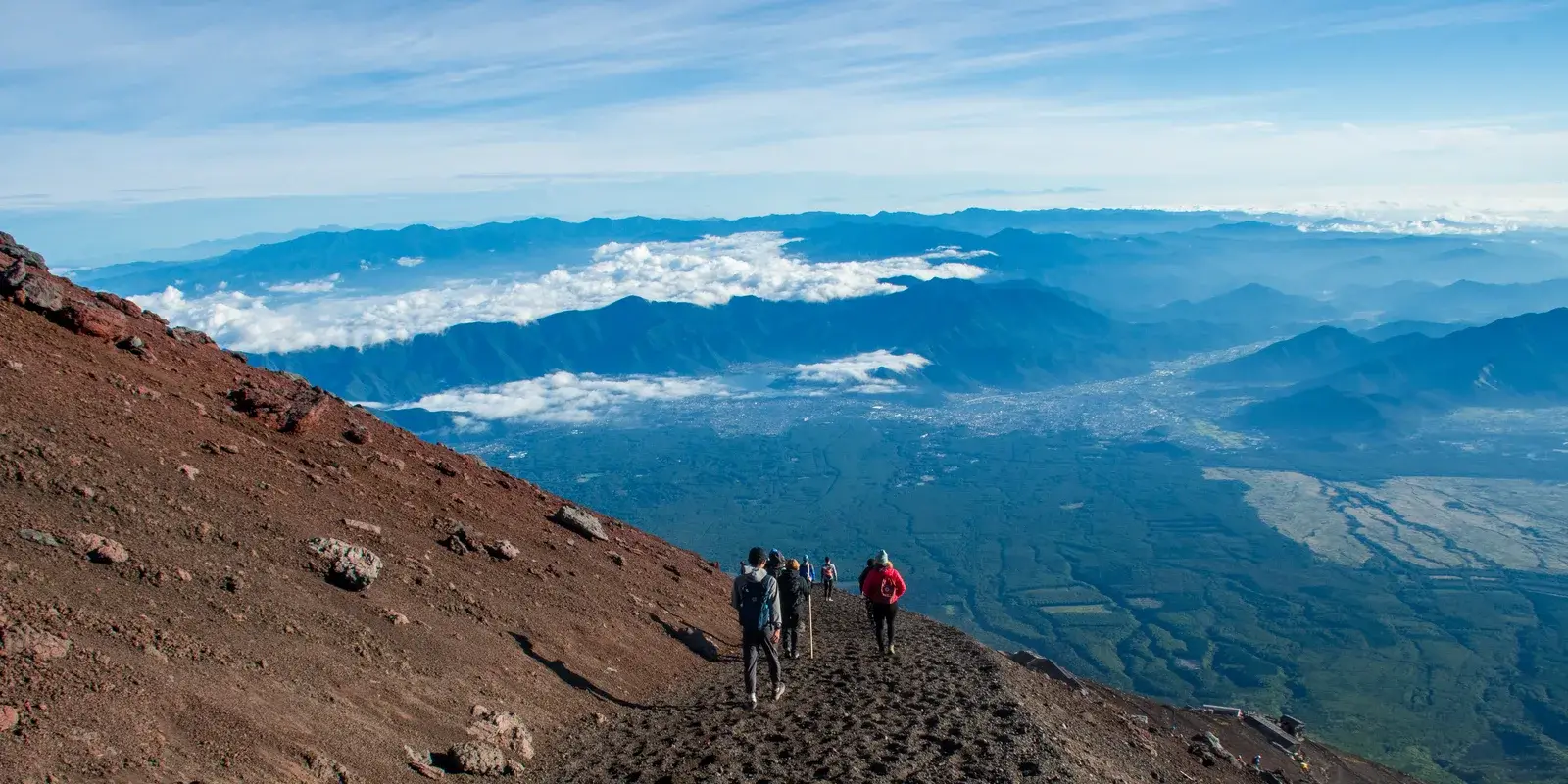 Climbers descending the Yoshida Trail on Mount Fuji's volcanic slope, with a sea of clouds and distant mountains below at dawn