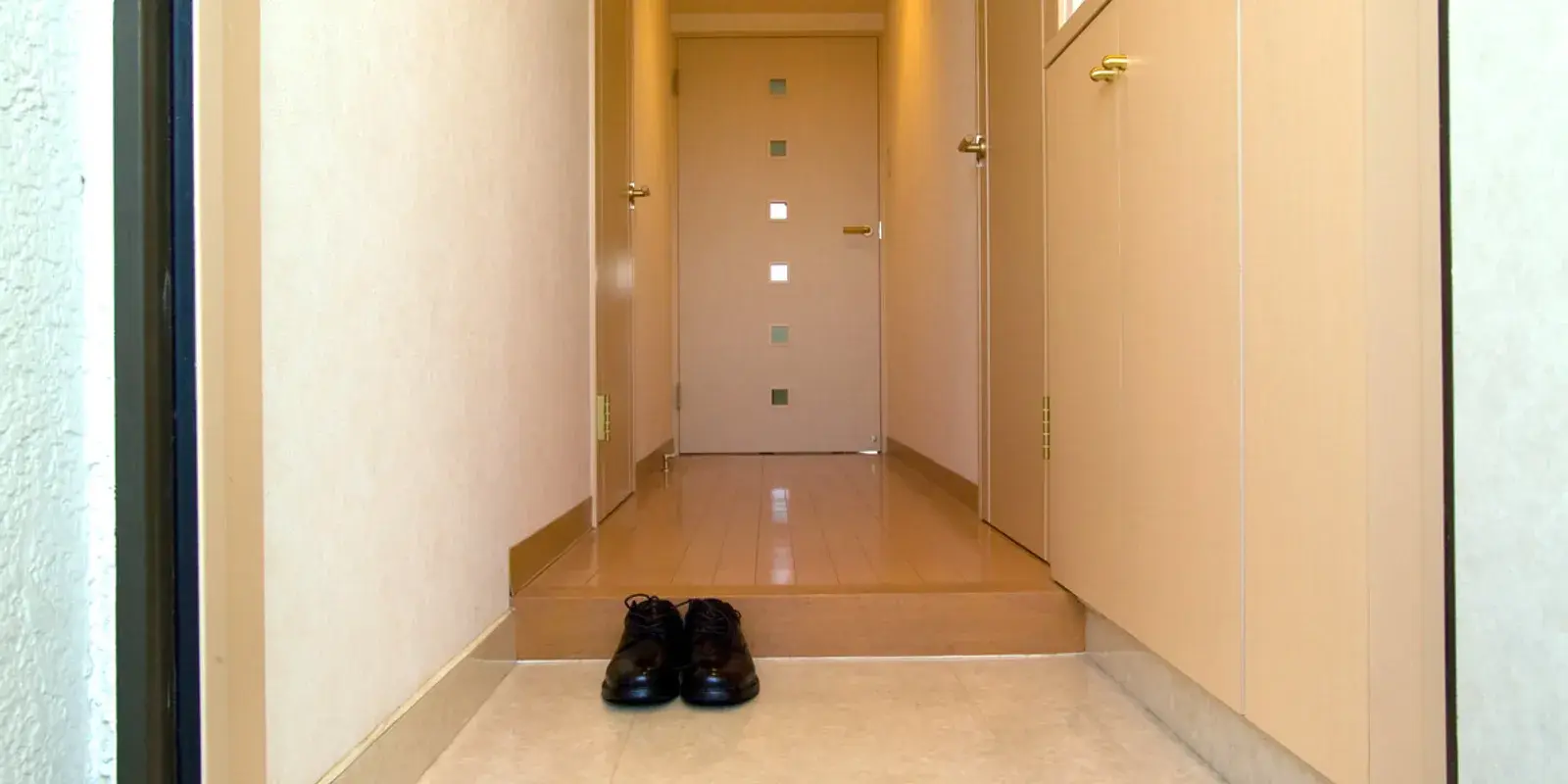 A single neatly placed pair of brown leather shoes on the tiled floor of a Japanese genkan entrance, with a shoe cabinet and a raised wooden hallway leading inside