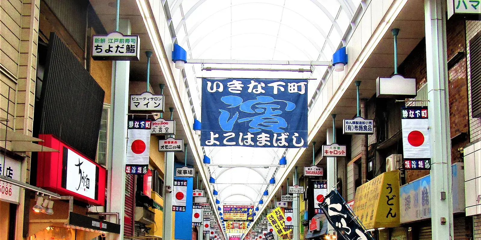 A covered Japanese shopping arcade (shotengai) lined with shops, signs, and banners, with the marked-price goods on display