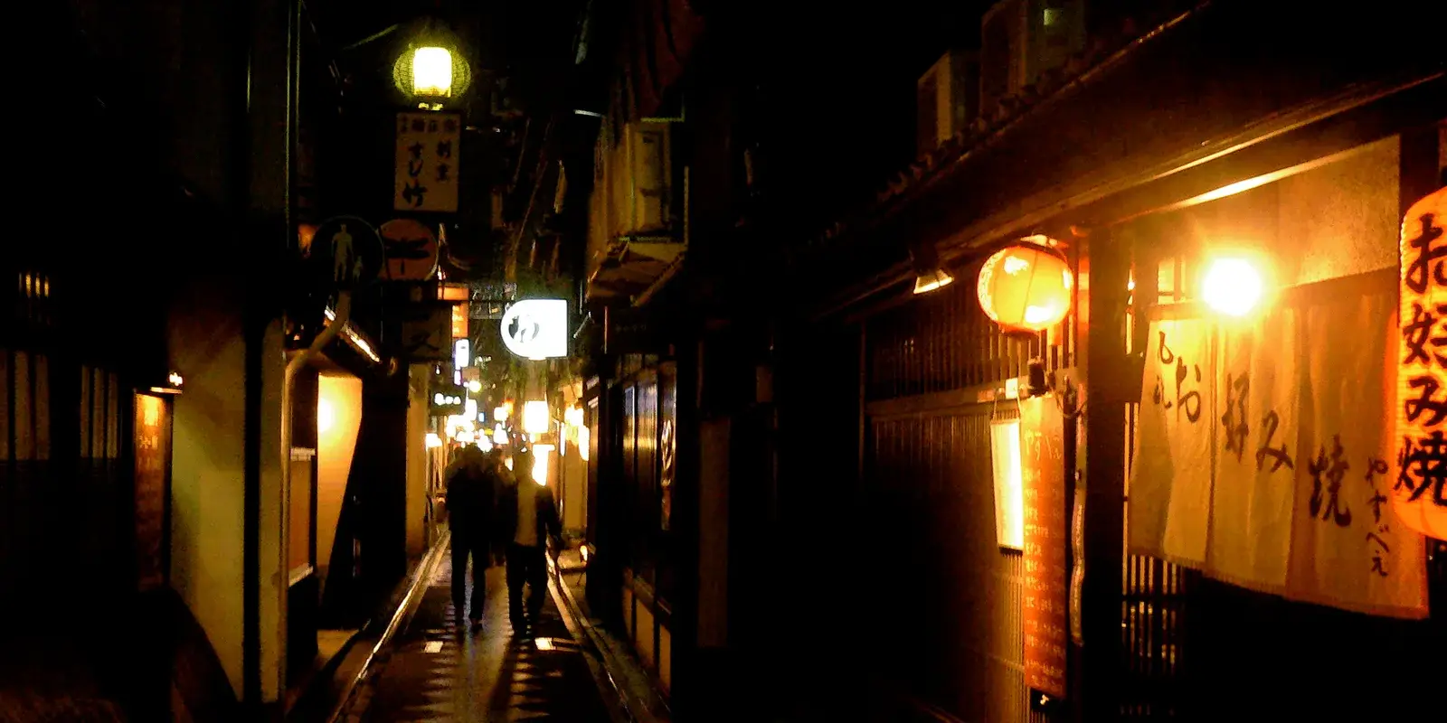 A warm, lantern-lit narrow eatery alley in Pontocho, Kyoto at night, with an okonomiyaki restaurant's glowing noren curtain on the right and two distant figures walking away