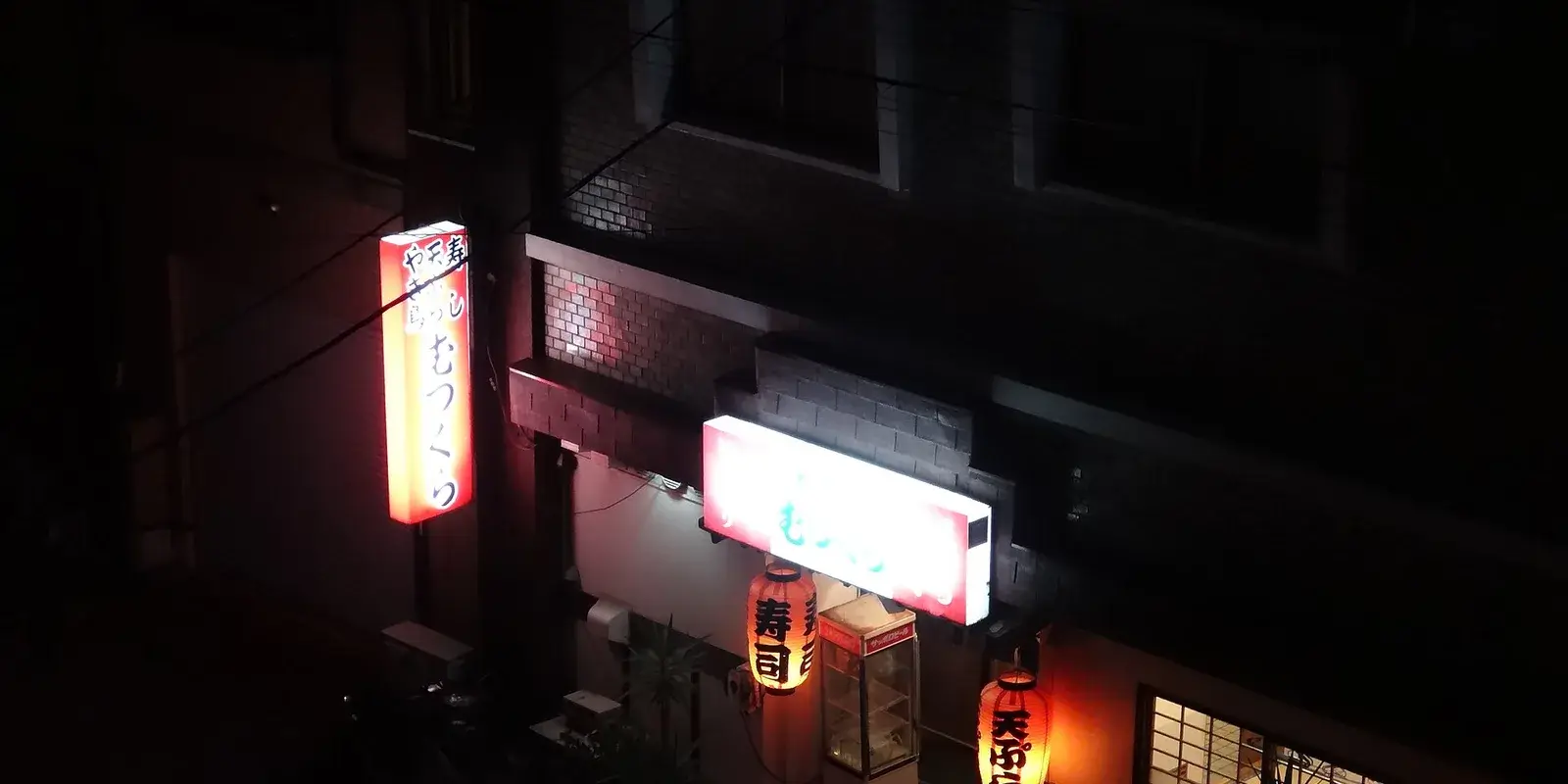 An izakaya storefront glowing at night, with red paper lanterns and an illuminated sushi and tempura sign above the entrance