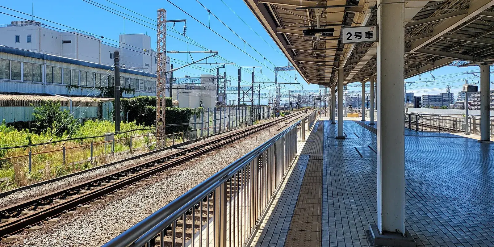 A calm, empty Shinkansen platform under a sunny blue sky, with tracks, the station canopy, and a "car 2" boarding sign — no people in sight
