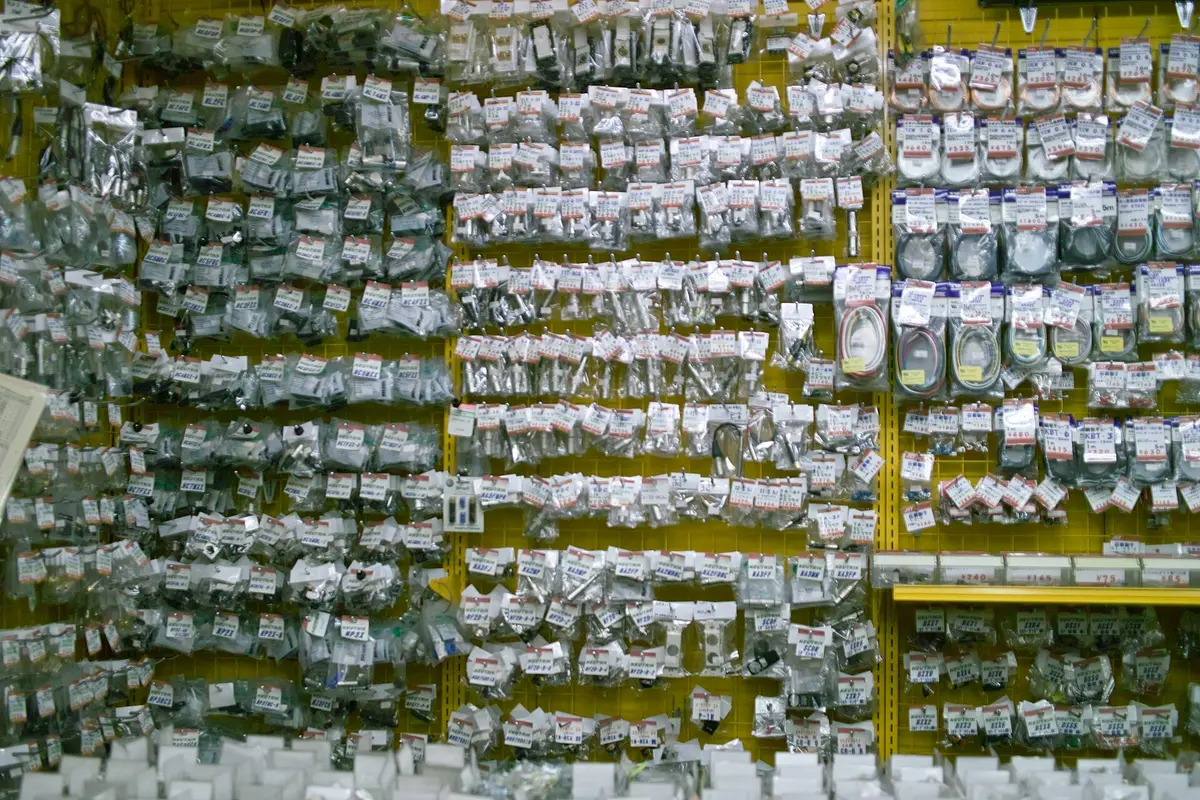 Rows of small bagged electronic connectors and cables hanging on a yellow pegboard wall in an Akihabara parts shop, each with its own printed price tag