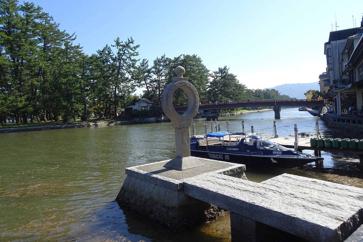 Die rote Drehbrücke und das steinerne Chie-no-wa-Ringdenkmal am Eingang zur Amanohashidate-Sandbank auf der Monju-Seite, mit einem Ausflugsboot, das daneben anlegt