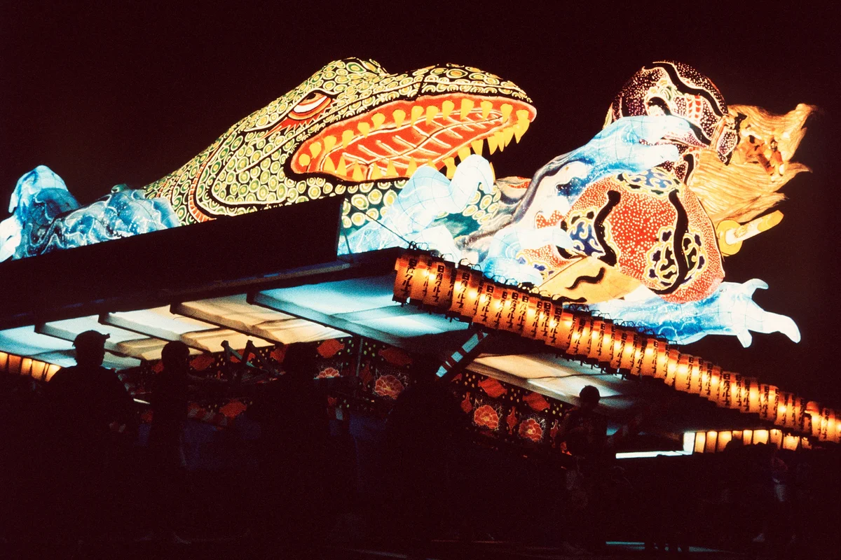 Silhouettes of the crowd watching an illuminated Nebuta float pass overhead at night
