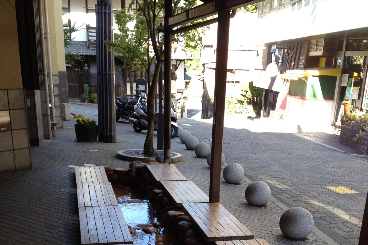 Holzbänke über einer flachen Rinne mit rötlichem Thermalwasser im kostenlosen Fußbad von Arima Onsen, neben einer abschüssigen Straße