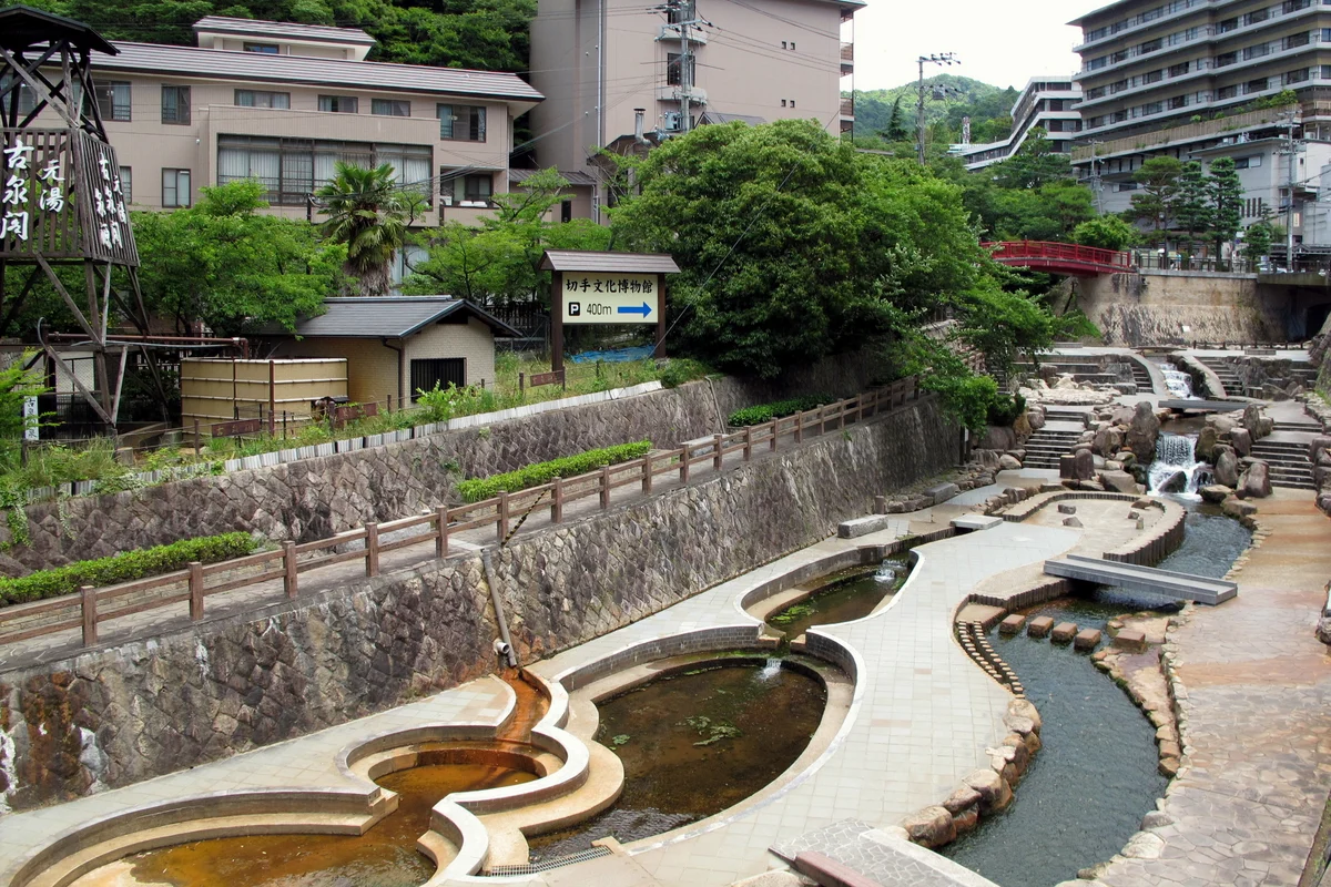 Becken mit Steineinfassung und Stufen am Arima-Fluss mitten im Onsen-Ort, dahinter Hotels am Hang und flussaufwärts eine rote Fußgängerbrücke