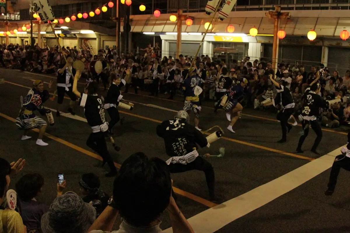 Awa Odori dancers in black happi coats moving in a low, wide stance down a Tokushima street closed to traffic, with people sitting along both kerbs