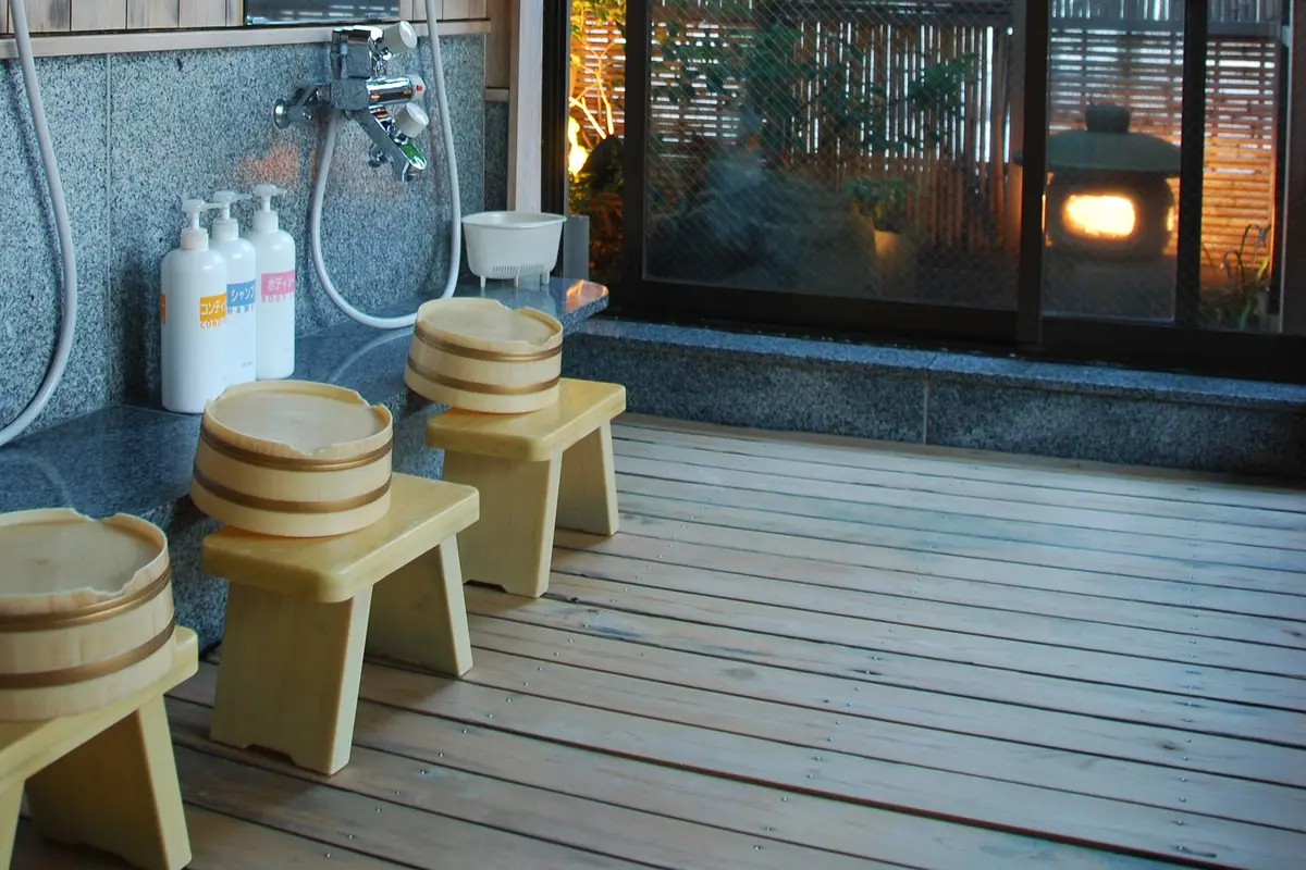 Three low wooden stools with wooden basins beside a handheld shower and tap at the washing station of a Japanese communal bath