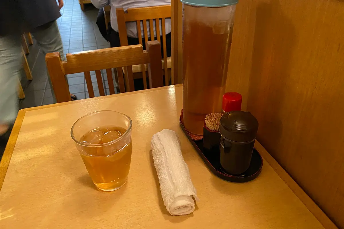 A glass of cold tea and a rolled white hand towel on a wooden table in a Japanese restaurant, next to a pitcher of tea, soy sauce and toothpicks