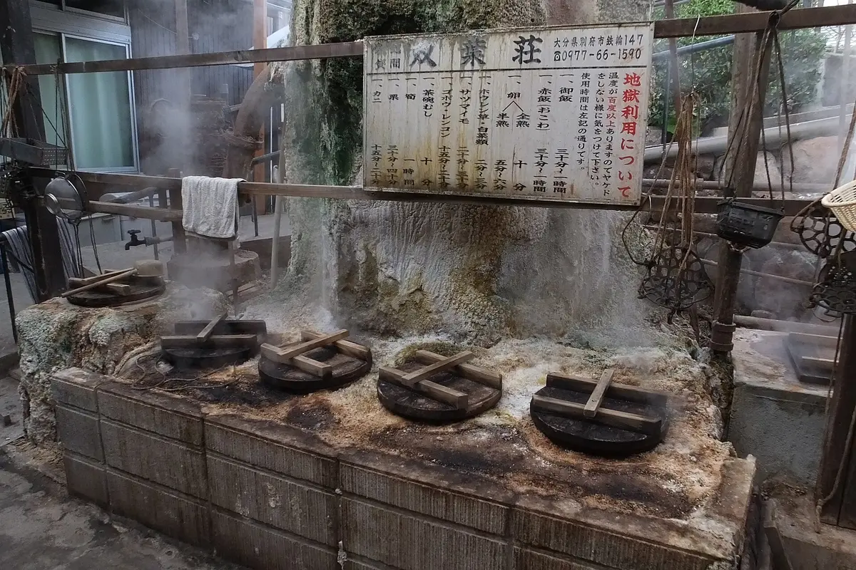 A row of stone steam vents with wooden lids in Kannawa, Beppu, steam escaping around the edges, under a sign listing steaming times