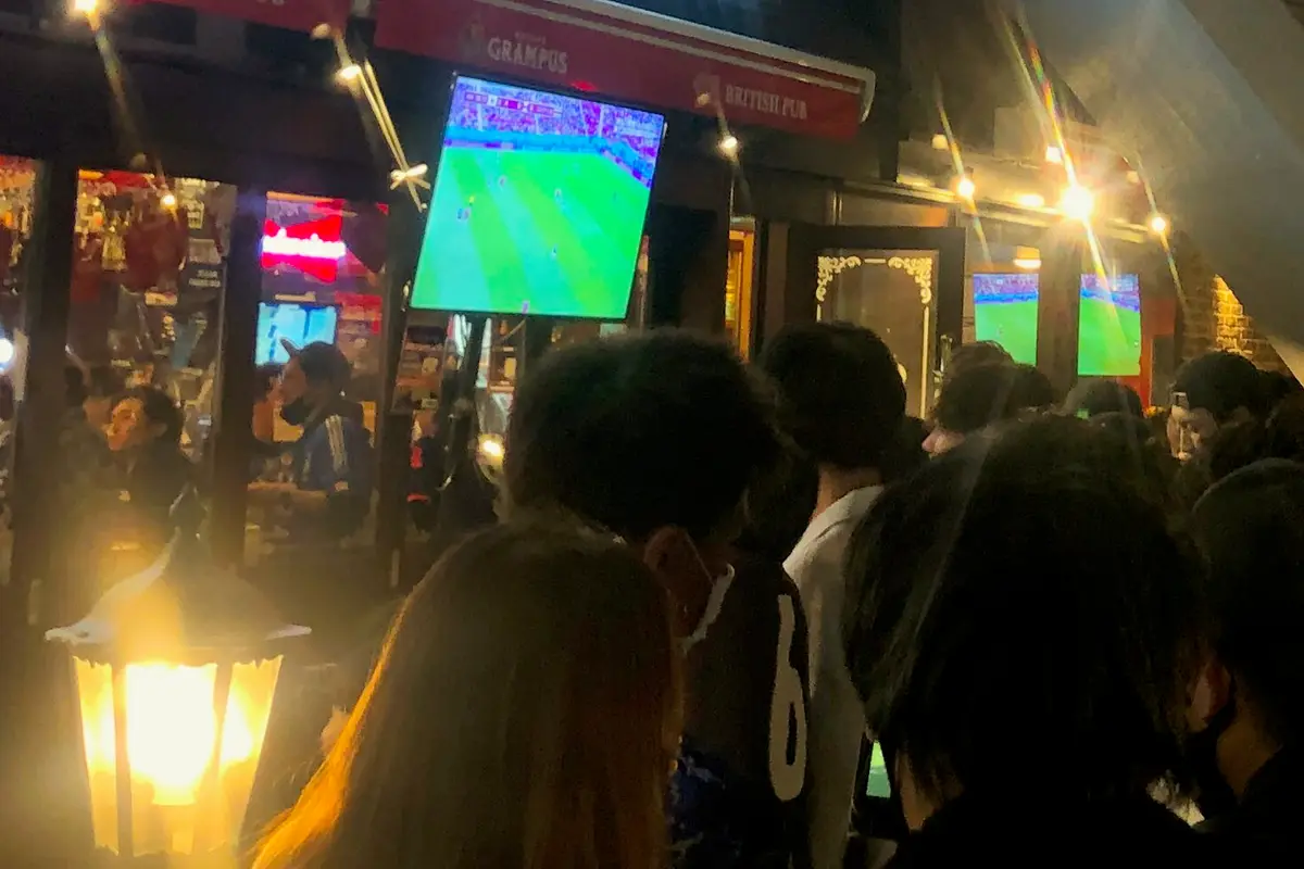 밤에 일본의 영국식 스포츠 펍 앞에 서서 대형 야외 스크린으로 축구 경기를 보는 사람들의 뒷모습