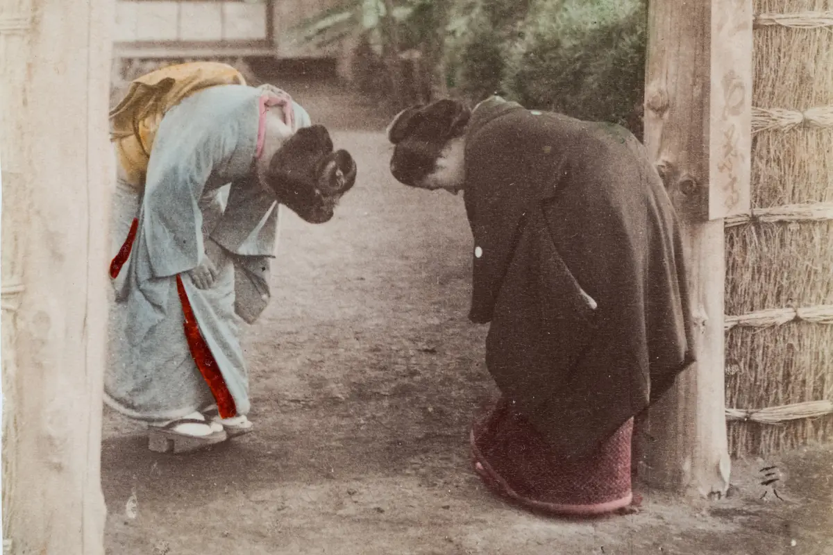 Two women in kimono bowing to each other under a wooden gateway, both with their heads lowered, in a hand-colored photograph from around 1880