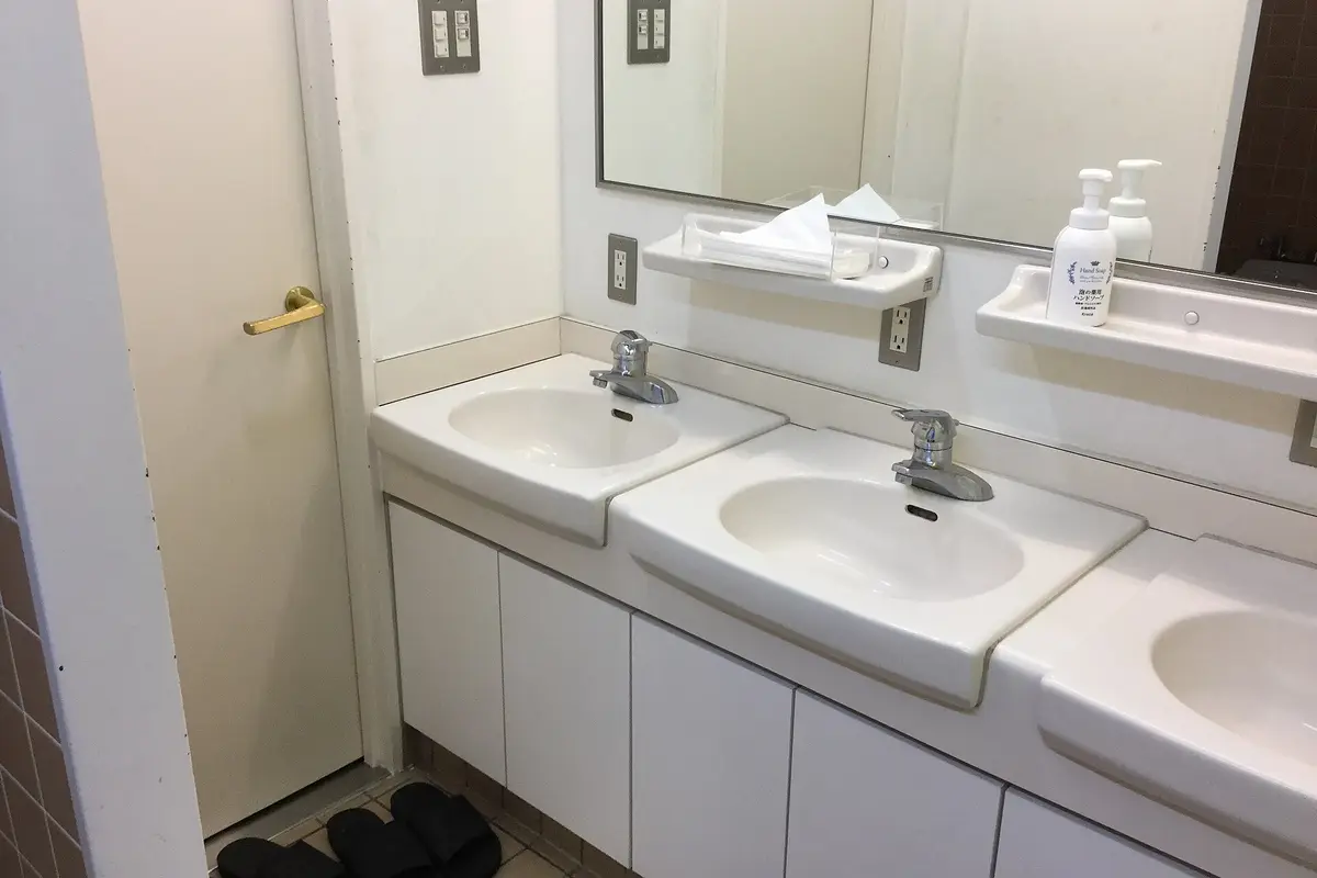 A row of shared washbasins with mirrors, hand soap and tissues, with house slippers on the tiled floor