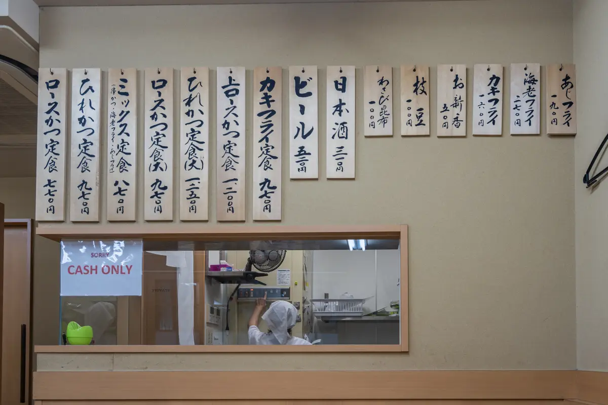 A printed notice reading SORRY CASH ONLY in the window of a small restaurant, below a row of hand-lettered wooden menu tags