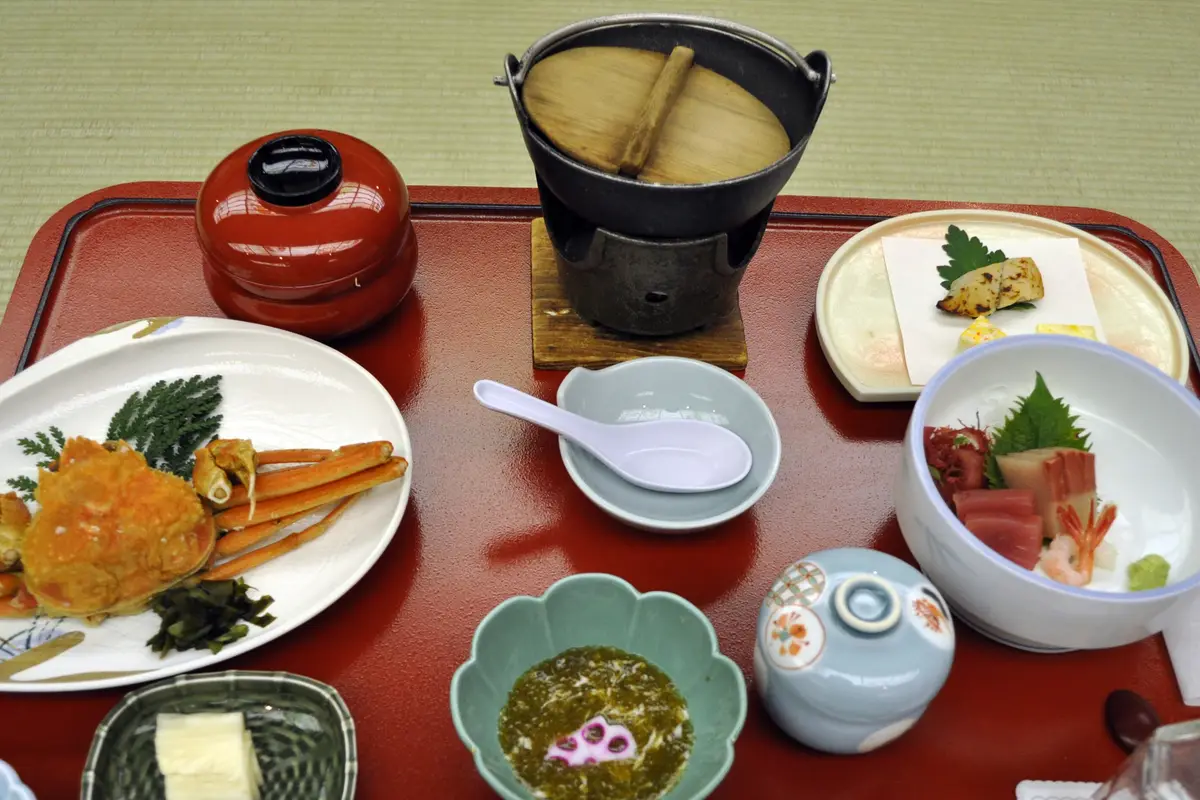 A ryokan dinner seen from above on a red lacquered tray set on tatami, with a plate of crab, a bowl of sashimi, a small iron pot on its own burner, and several small covered bowls
