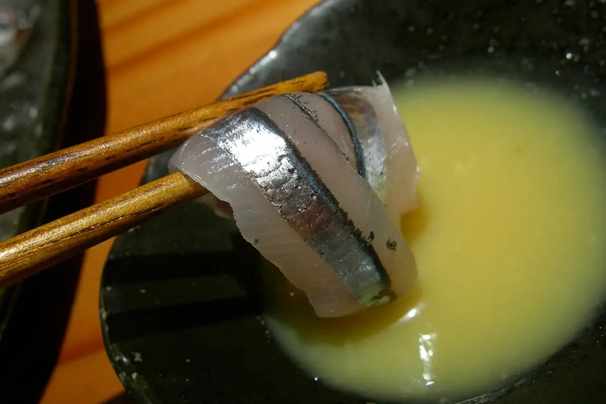 Close view of the tapered tips of a pair of wooden chopsticks holding a slice of raw fish above a dish of sauce