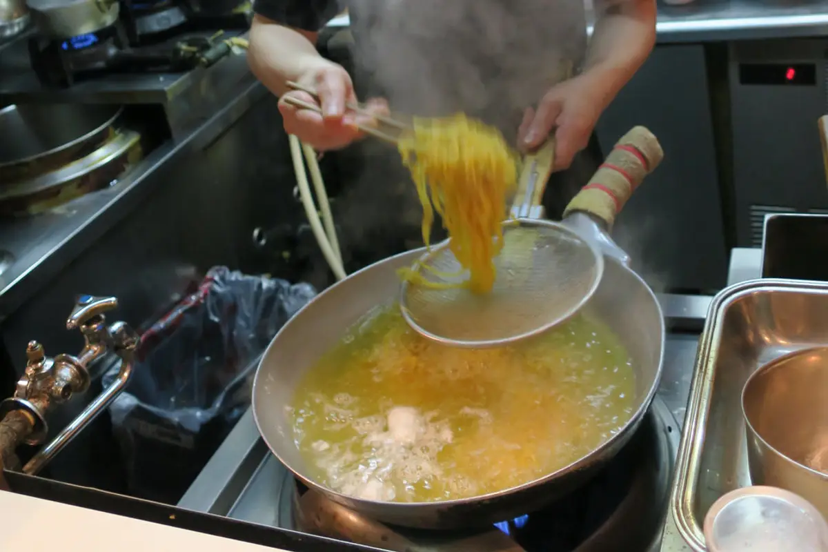 Hands lifting a basket of noodles from a boiling pot, seen from a seat at the counter of a small ramen shop