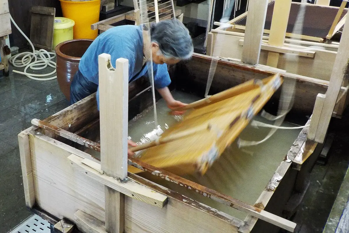 A person bent over a vat, drawing a bamboo screen through the pulp to form a sheet of handmade paper