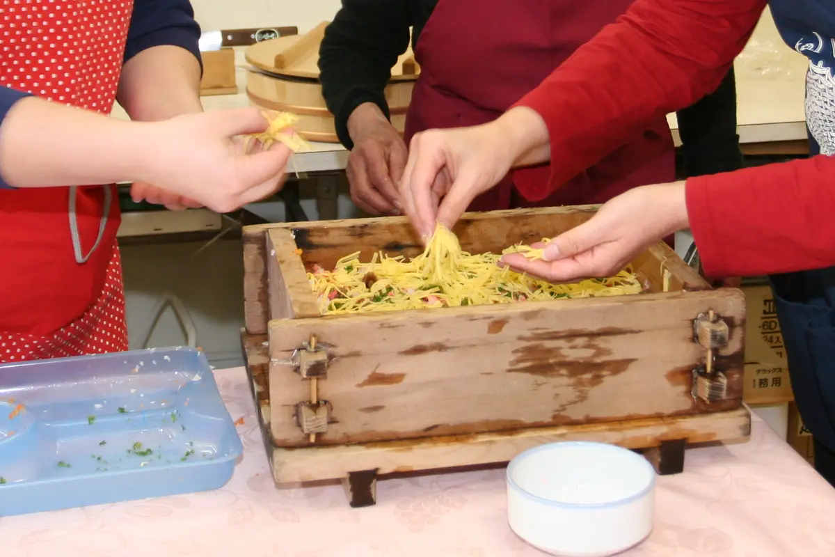 Hände streuen Eierstreifen über eine Schicht Sushireis in einer hölzernen Pressform, in einem Kochkurs in Japan