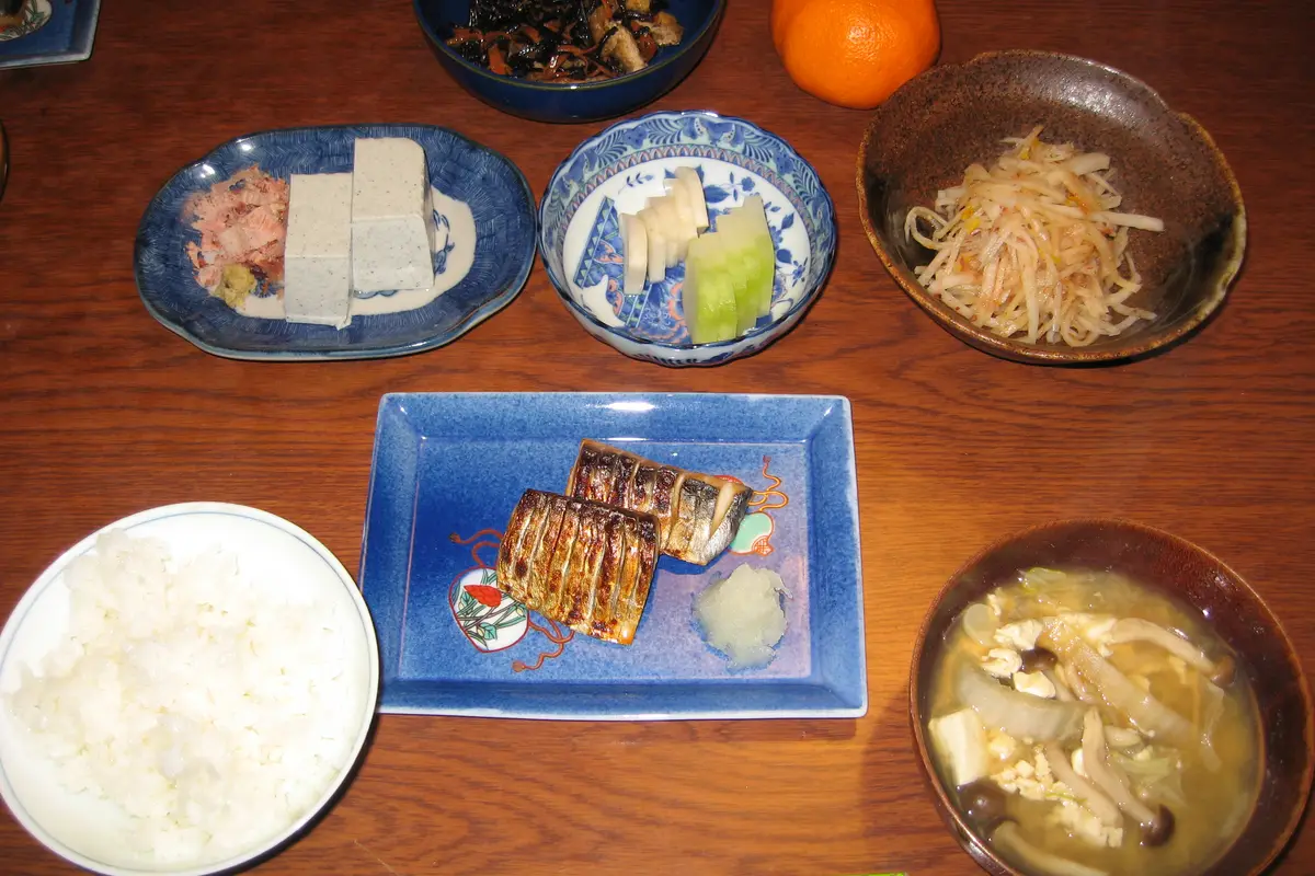 Ein alltägliches Abendessen in einem japanischen Zuhause von oben gesehen: eine Schale Reis, Misosuppe, gegrillter Fisch, kalter Tofu, eingelegtes Gemüse und ein Sojasprossen-Salat auf einem Holztisch