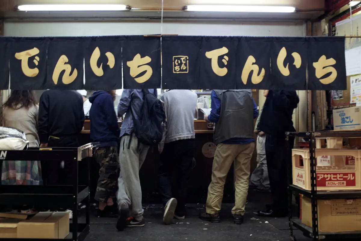 Customers standing shoulder to shoulder at the counter of a small standing bar, seen from behind beneath a navy blue noren curtain