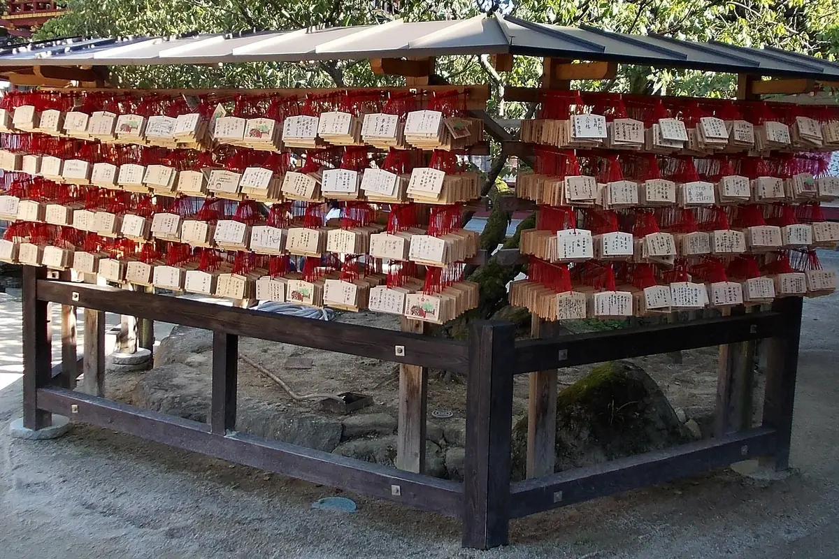Rows of small wooden ema plaques tied with red cords, hanging on a covered rack in the grounds of Dazaifu Tenmangu