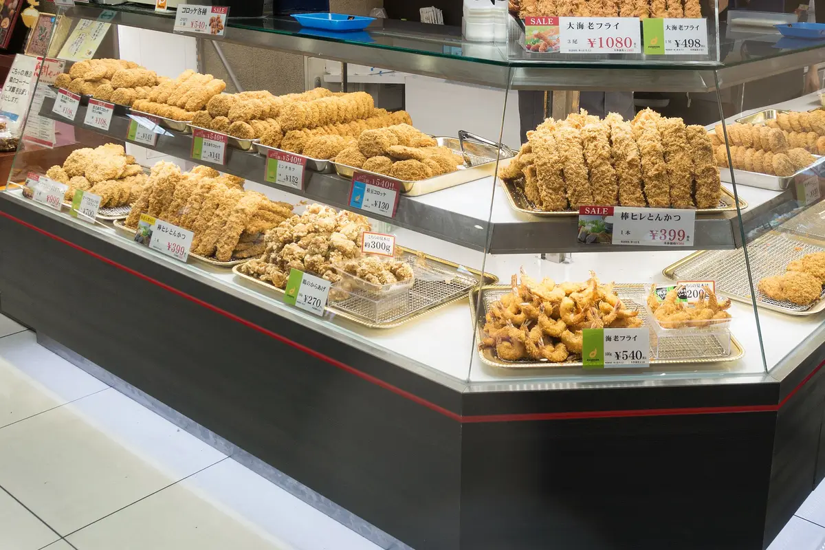 An open deli case of croquettes, pork cutlets and fried prawns, with a small printed card beside each tray giving the price by weight or by the piece