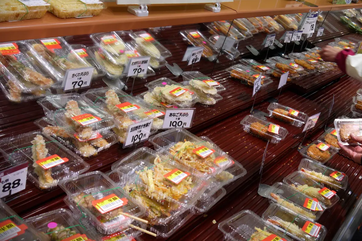 Trays of packed tempura and fried side dishes on a display shelf, most of them carrying a red half-price label, with a hand reaching in from the right