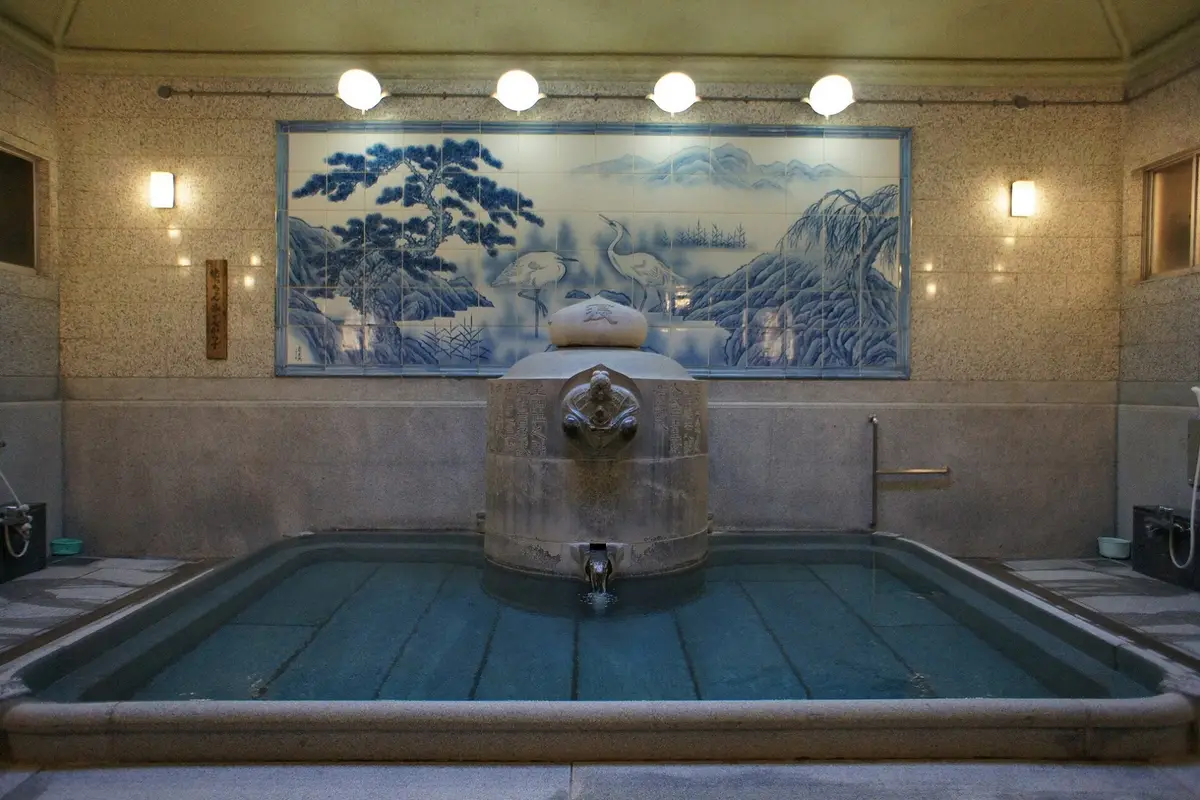 Inside the Dogo Onsen Honkan: a single rectangular stone bath with hot water pouring from the carved stone pillar in its middle, below a blue and white tile panel of herons and pines
