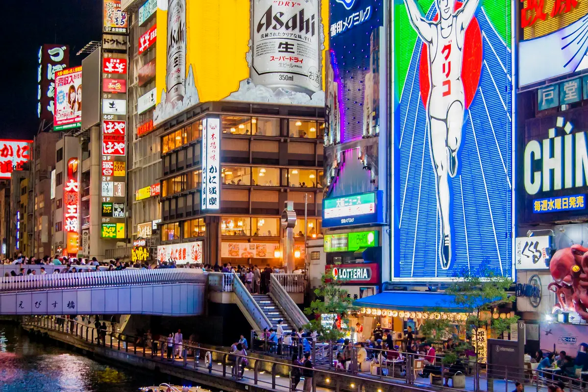 El puente Ebisubashi lleno de gente por la noche, bajo un muro de enormes carteles iluminados sobre el canal de Dotonbori