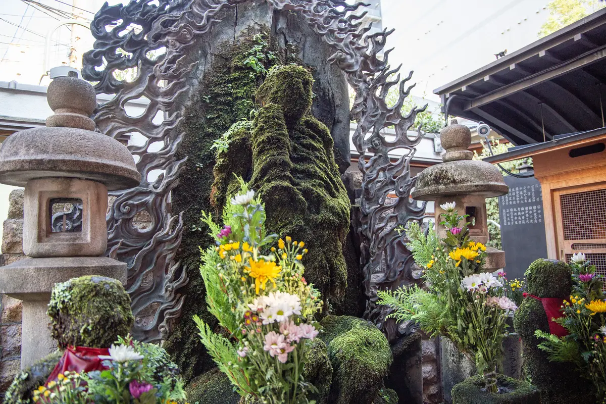 Una estatua de piedra cubierta de musgo entre dos linternas de piedra, con flores frescas a sus pies, en Hozenji Yokocho