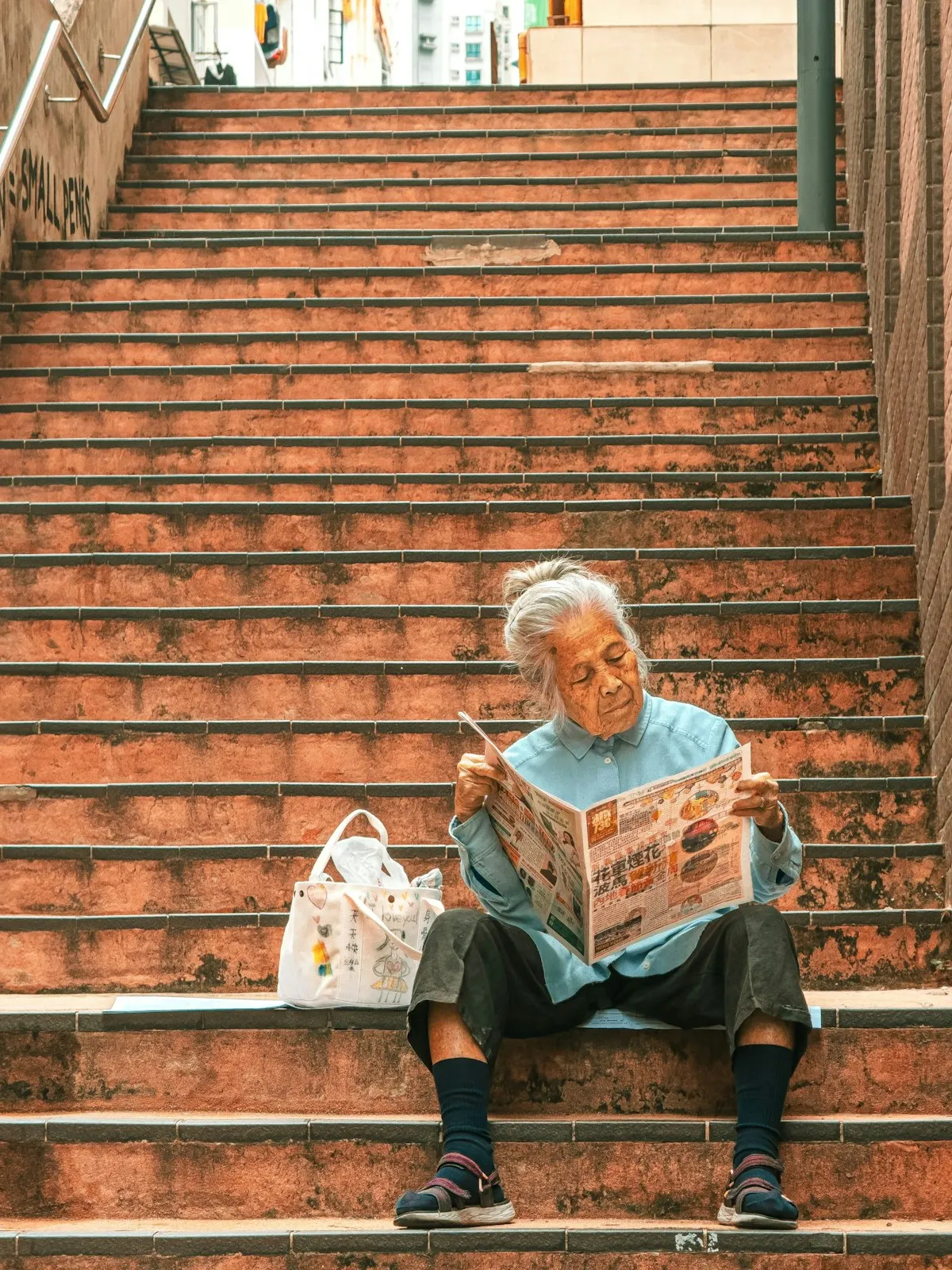 An elderly Japanese woman sitting on outdoor stairs reading a newspaper