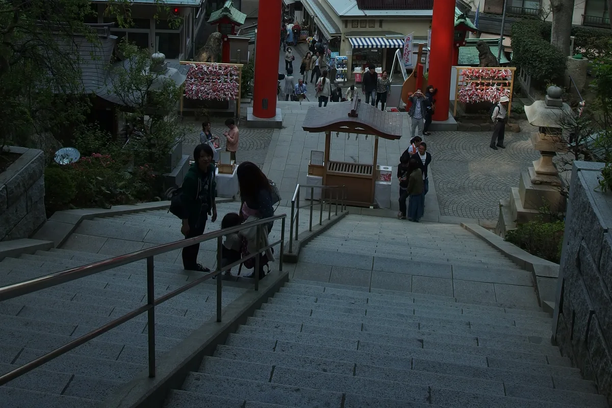 A red torii gate at the top of a long stone staircase on Enoshima, with visitors climbing and shops lining the approach