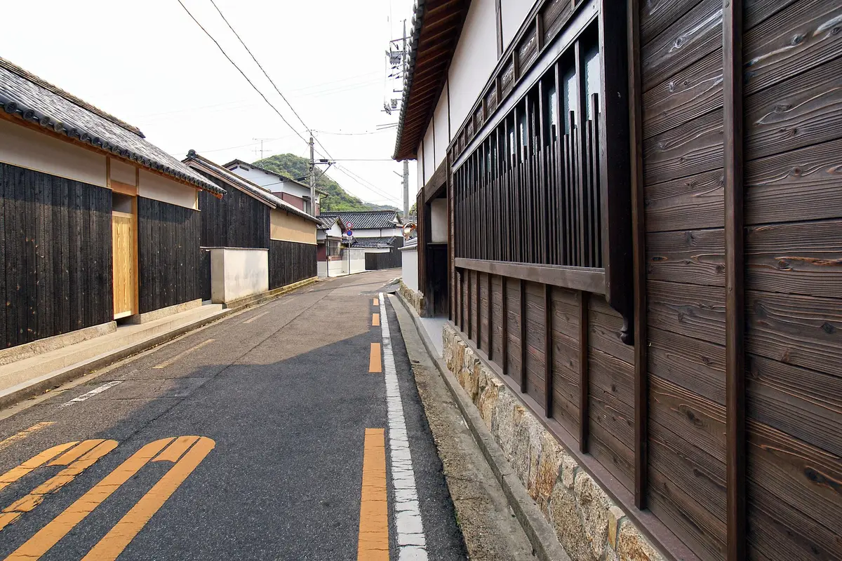 A narrow residential lane on the island of Naoshima, lined with old houses of dark charred cedar boards, white plaster walls and wooden lattice windows, with no one on the road