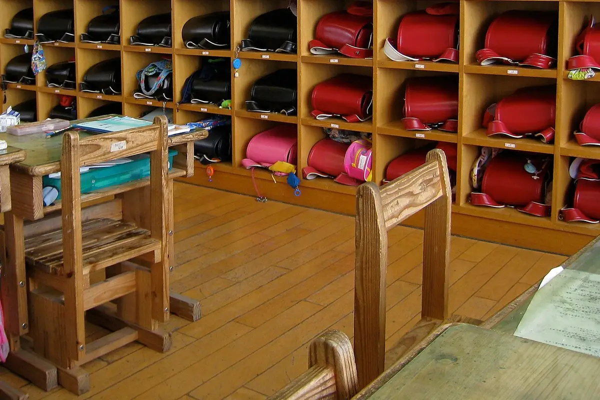 Rows of wooden cubbies in a Japanese elementary school classroom, each holding a black, red or pink randoseru backpack, with small desks and chairs on the wooden floor in front